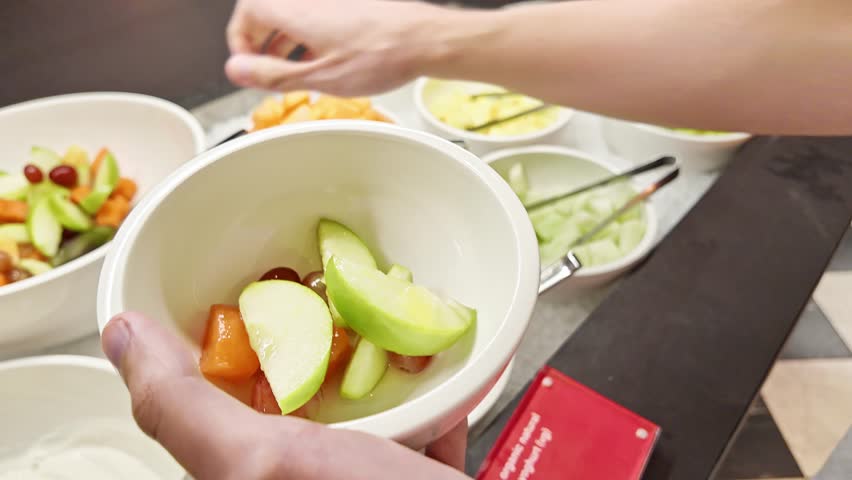 Man fills bowl with sliced melon and green apple at hotel breakfast buffet, enjoying healthy start to the day, hotel breakfast, healthy eating