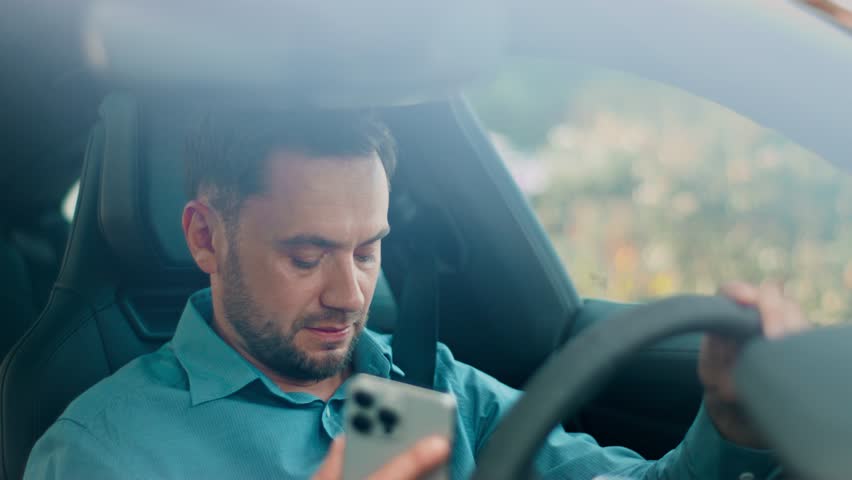 Annoyed man holding phone in one hand while raising clenched hand near face. Showing negative emotions after update. Reacting to difficult news while sitting in drivers seat with tense expression.