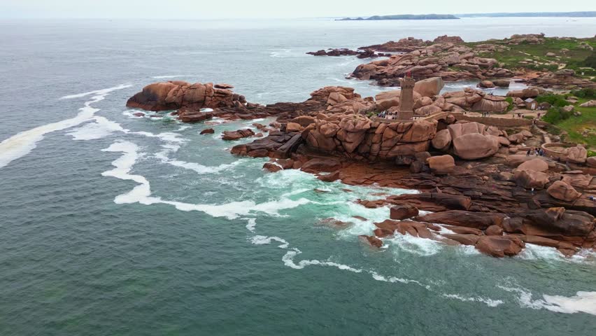 Approaching panoramic drone movement the rocky Côte de Granit Rose with the Men Ruz lighthouse and visitors, Ploumanac'h, Brittany, France.