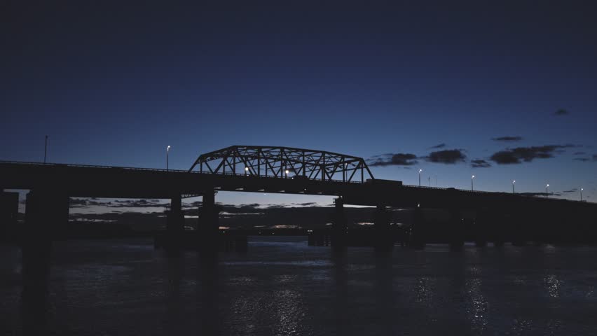 Timelapse of a semi-truck crossing a bridge at dusk under dramatic clouds. Reflections ripple below on a calm river.