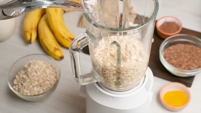 Clean water poured from a carafe into a glass blender with whole oat flakes. Mixing oats and water creates oat milk a vegan, vegetarian base for smoothies and plant-based cocktails without animal fat