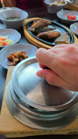 A man's hand opens a steaming hot metal pot to reveal freshly cooked rice with beans and chestnut, served in a Korean traditional restaurant with assorted side dishes and fried fish - vertical