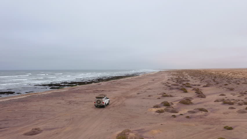 Drone follows a lone vehicle driving on a vast, empty beach near Swakopmund on a moody, overcast day