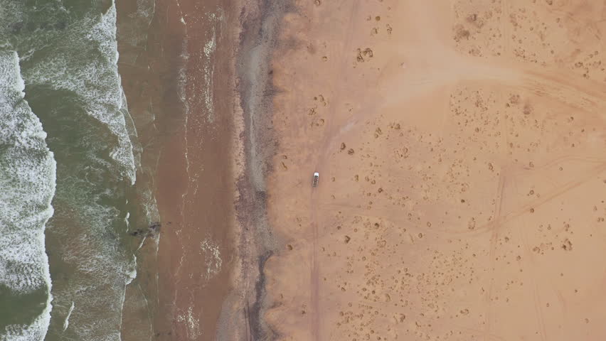 Top down drone shot of an off-road vehicle parked alone on a wide sandy beach near Swakopmund in Namibia, with soft waves rolling in under cloudy skies