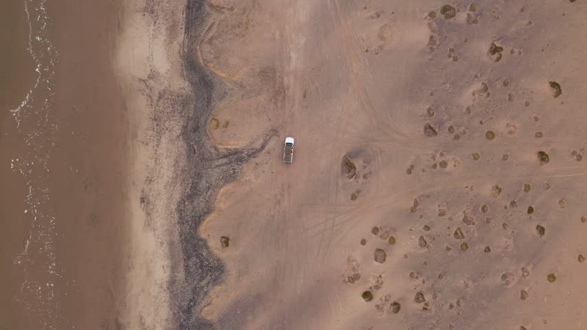 Top down drone footage following an off-road vehicle driving along the sandy beach near Swakopmund, Namibia, under overcast skies