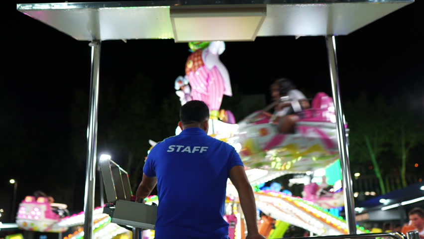 Man in blue staff shirt operating amusement park ride at night filmed from behind