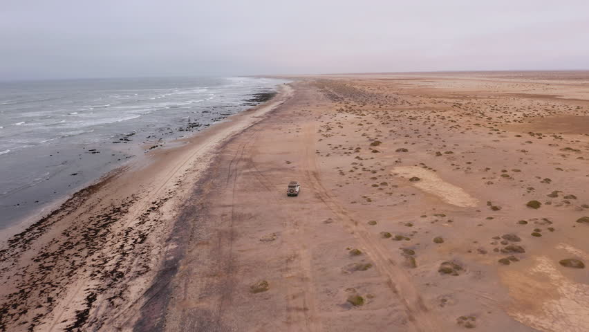 Aerial: Following of an off-road vehicle cruising along the wide sandy beach near Swakopmund, Namibia, under a grey overcast sky