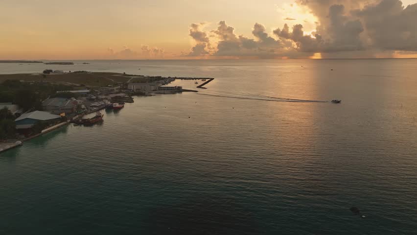 Aerial view of Thulusdhoo Island where the buildings meet the turquoise ocean, boats are docked, and the sun sets behind the clouds, Thulusdhoo, Kaafu Atoll, Maldives.