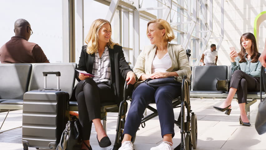 Businesswoman Sitting In Airport Departure With Female Colleague In Wheelchair Talking Together