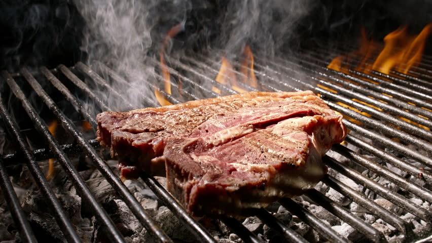 Slow motion video in 4K of T-bone steak cooking on barbecue grill with flames, smoke, and seasonings. Close-up footage for culinary, restaurant, and barbecue themes.