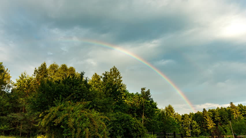 Rainbow over summer forest and meadow. Beautiful dramatic sky timelapse.