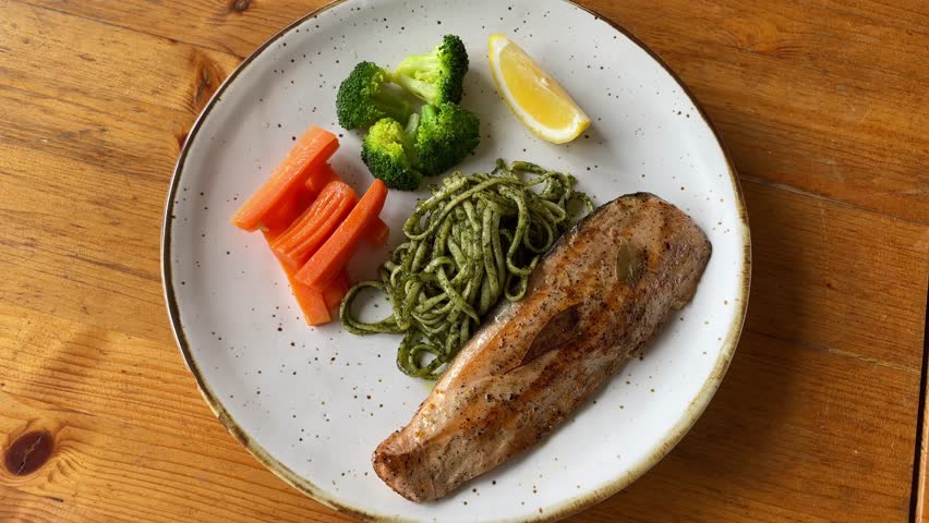 Grilled fish fillet served with steamed broccoli, carrots, green pesto pasta, and a lemon wedge on a rustic ceramic plate over a wooden table, healthy balanced meal concept