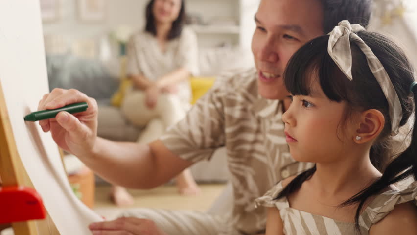 Happy Asian family spending quality time together at home. Father and daughter drawing on the floor with colored pens while mother works on a digital tablet smartphone on the sofa. Warm, modern living