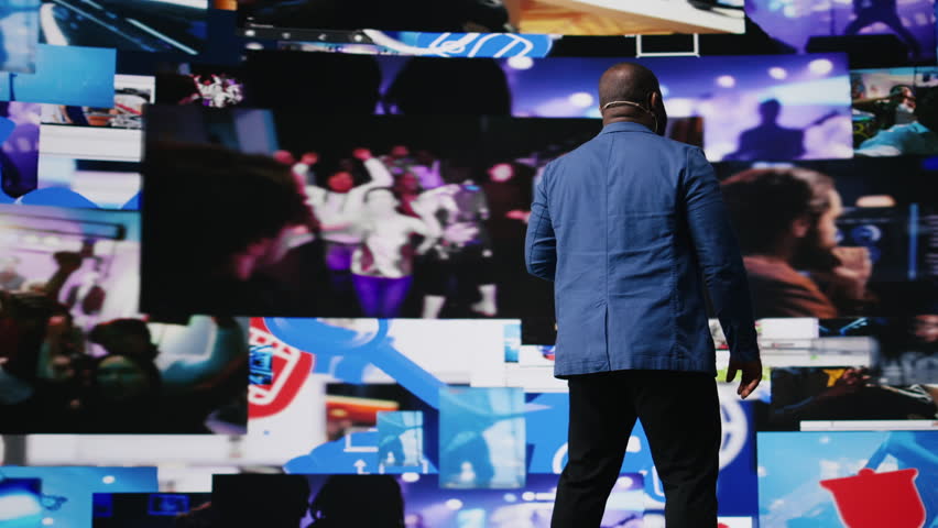 Black presenter hosting conference on stage surrounded by media visuals, symbolizing global convention with live audience to discuss about online trends and mobile apps. Public event. Camera A.