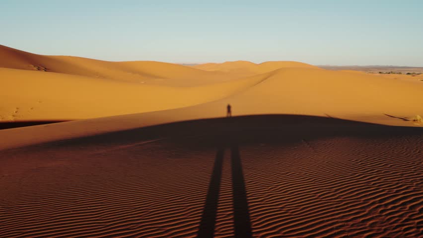 Vast expanse of the Sahara Desert with beautiful, rolling sand dunes at sunrise. A lone figure's shadow stretches across the dunes, beauty of this Moroccan landscape.