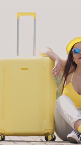 A cheerful young woman in stylish summer attire sits at a busy train station with her bright yellow suitcase, eagerly looking forward to an exciting vacation full of adventure and happiness