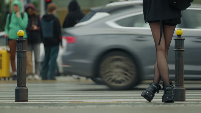 Close-up of young slim woman's legs in black skirt and tights, wearing edgy black boots, standing at city crosswalk as cars pass in slow motion, urban street scene, daylight cloudy summer day