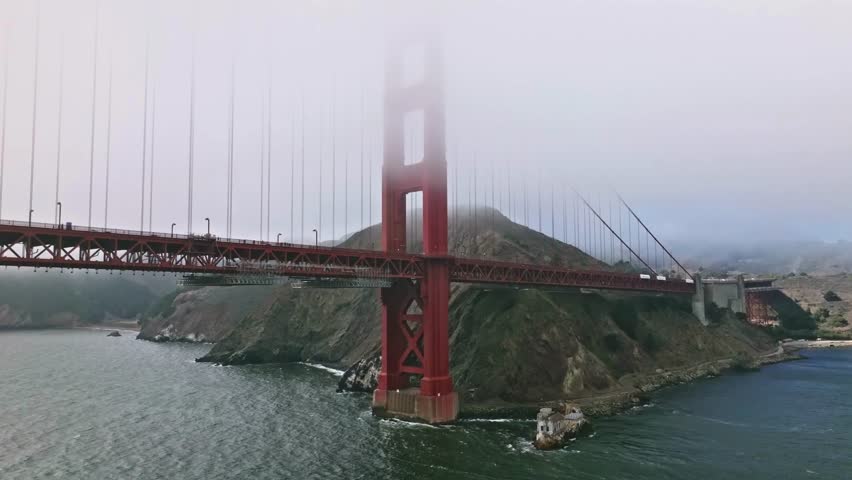 Fog envelops iconic Golden Gate Bridge as dawn breaks over San Francisco Bay. Calm waters reflect structures while fog adds mystique to morning skyline.
