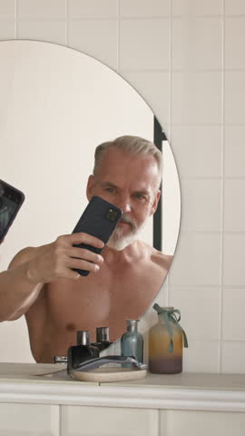 Vertical shot of shirtless senior man with smartphone standing in front of round mirror hanging on wall of bathroom and looking at himself on screen of gadget while taking selfie