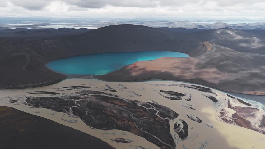 Aerial view of a vibrant blue crater lake surrounded by dark volcanic terrain, with rivers, rugged slopes, and distant mountains under a cloudy sky.