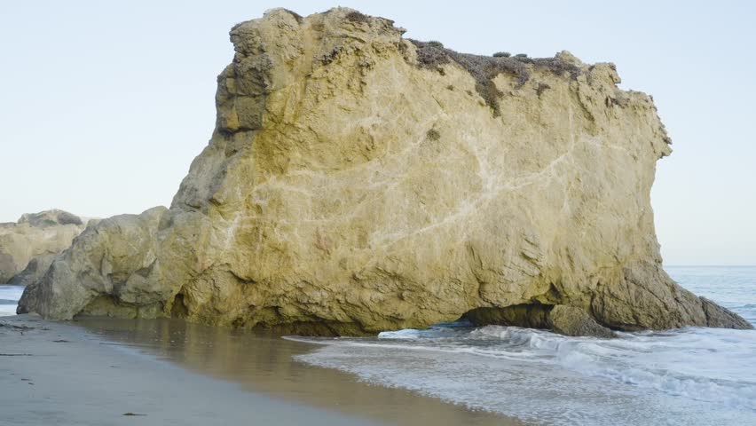 Ocean waves wash against a large rock formation at the shoreline under soft daylight on a calm, empty beach.