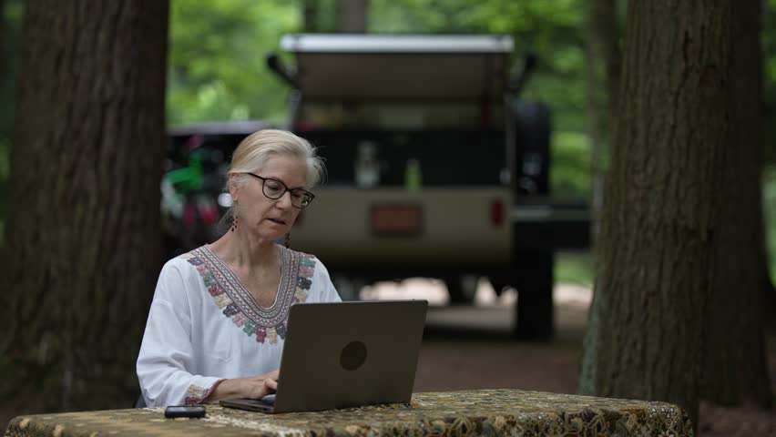 A mature woman engages with her computer and smartphone while seated at a table in a forested campground, surrounded by trees and her teardrop camper trailer.