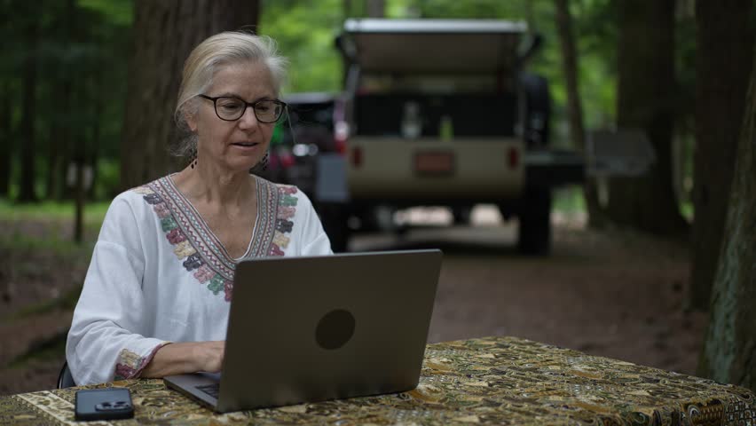 A retired woman happily uses her laptop and smartphone at a campground, with her teardrop camper trailer in the background surrounded by trees.