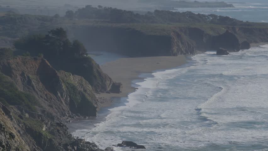 Aerial view of dramatic cliffs, sandy beaches, and powerful ocean waves rolling onto a rocky California shoreline under soft sunlight.