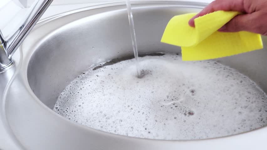 A hand gently presses a yellow sponge under running water in a sink full of suds-an intimate glimpse into daily rhythm, where cleanliness meets quiet focus