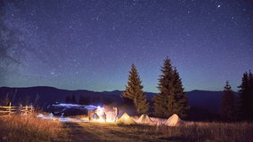 Night camping in mountains. Amidst forested mountain landscape, two tents glow warmly beside campfire under brilliant, star-filled sky. Silhouettes of tourists resting at campsite under Milky Way. - Powered by Shutterstock - Get 15% off with code: PIKWIZARD15