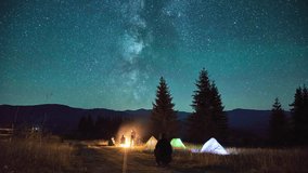 Night camping in mountains. Amidst forested mountain landscape, two tents glow warmly beside campfire under brilliant, star-filled sky. Silhouettes of tourists resting at campsite under Milky Way. - Powered by Shutterstock - Get 15% off with code: PIKWIZARD15