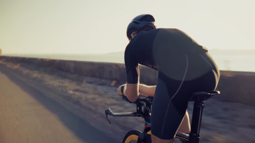 cyclist in aerodynamic black gear and helmet rides on open rural road. Cyclist in racing position. Endurance, speed, and professional outdoor cycling. Summer session cycling. Endurance sport bike ride