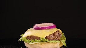 A sesame seed bun top is dropped onto a stacked beef burger with lettuce, cheese, tomato, and onion against a black background, using bright studio lighting and slow-motion capture - Powered by Shutterstock - Get 15% off with code: PIKWIZARD15