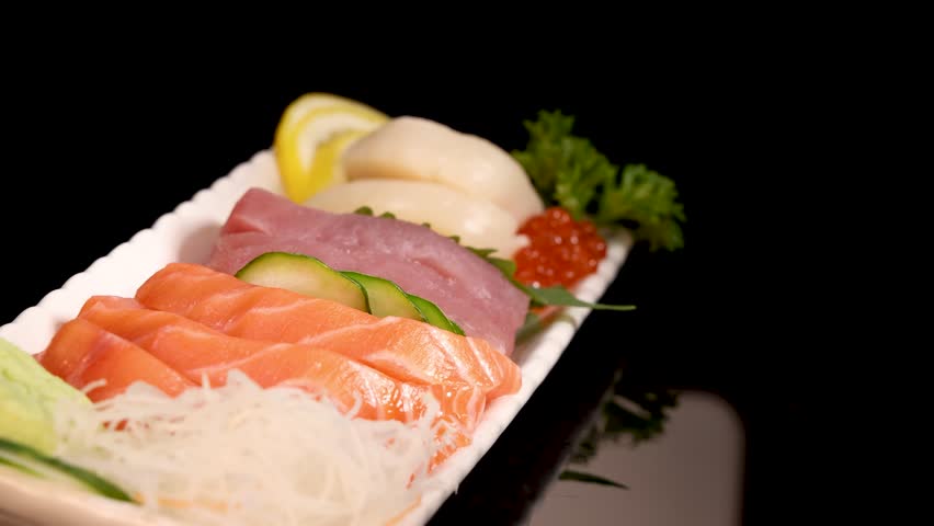 A pair of chopsticks selects salmon roe from an assorted sashimi sushi set on a white plate, under studio lighting with a black background