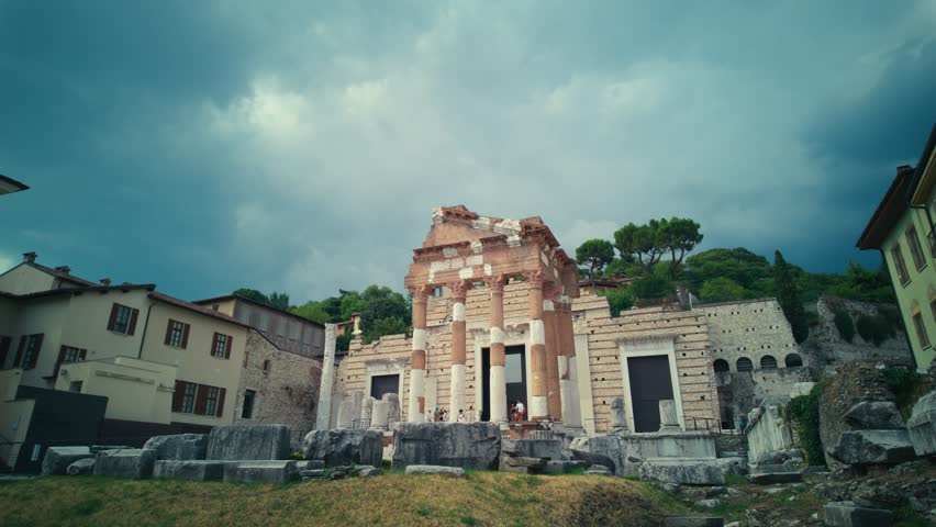 Timelapse. Ancient Roman Capitolium ruins in Brescia, Italy, under dramatic cloudy skies. Historical landmark surrounded by old houses and lush greenery.