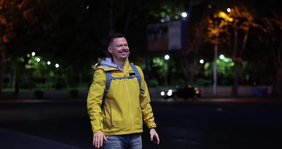 Man in yellow jacket waves hand high above head on evening street. Person jumps up and down smiling after noticing old friend on night street