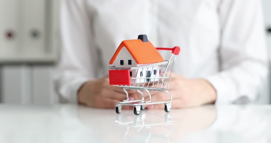 Woman hand pushes toy shopping card with plastic house at desk. Female realtor offers to buy country cottage on favorable terms in agency office