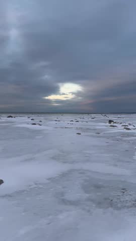 Frozen lakeshore under dramatic winter clouds at sunset, with snow and scattered ice formations captured in vertical format with subtle ambient audio.