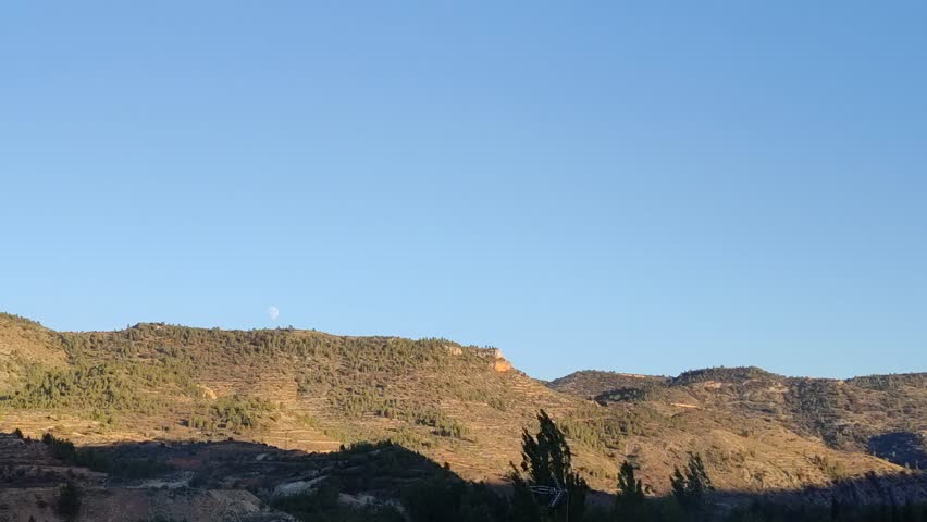 Timelapse of mountain landscape with clear sky and distant moon rising during the transition from sunset to night