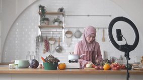 Muslim woman vlogger live streaming cooking online in kitchen look at camera with vegetables, content creator culinary class at home studio, halal food influencer cooking live, video for social media - Powered by Shutterstock - Get 15% off with code: PIKWIZARD15
