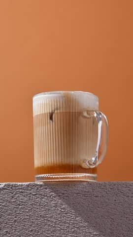 Hand stirs creamy iced autumn latte in clear glass against orange background, blending layers into smooth, inviting seasonal beverage.