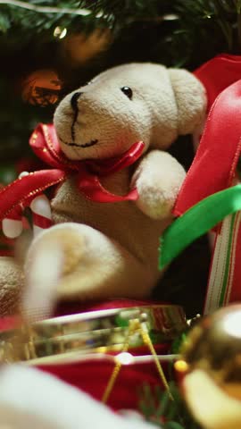 Soft teddy bear toy with red ribbon rotating among Christmas tree decorations in vertical composition