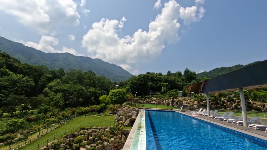A peaceful time-lapse captures clouds moving across a blue sky above a luxury infinity swimming pool with lounge chairs, overlooking a beautiful mountain landscape in Gapyeong, South Korea.