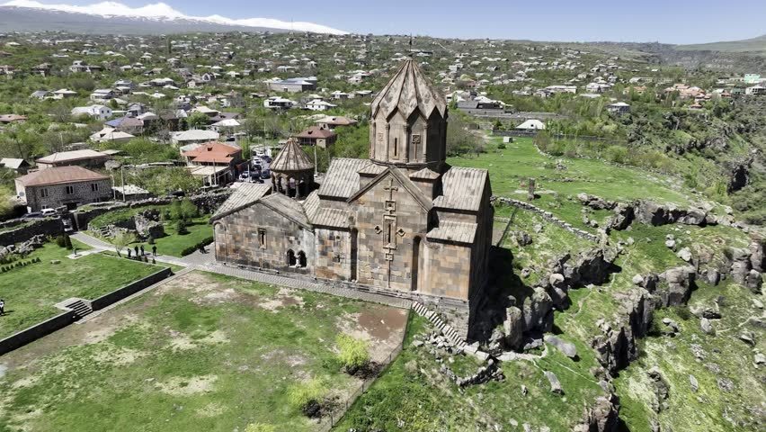 An aerial view captures the ancient Hovhannavank Monastery surrounded by green hills and a village on a sunny day in Ohanavan, Armenia.
