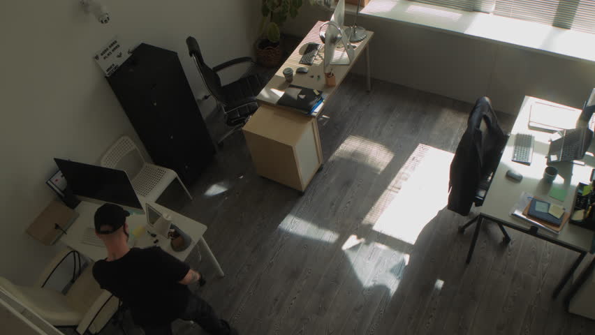 High angle shot of masked male criminal walking from desk to desk in office room searching for confidential corporate data looking through documents during industrial espionage