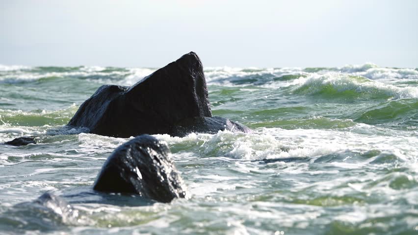 Sunlit sea waves crash against rugged coastal rocks. Dynamic motion captured in serene yet powerful seascape. Soft sky contrasts with churning water, evoking tranquility