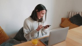 A young woman sits at her dining table, working remotely. She smiles while using her phone and laptop, with a glass of orange juice beside her. - Powered by Shutterstock - Get 15% off with code: PIKWIZARD15