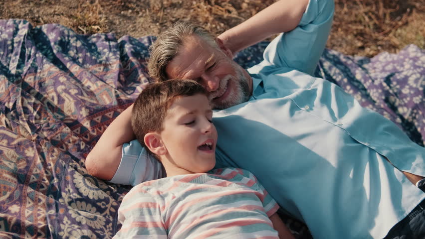 Grandfather and grandson relax together on a blanket outdoors. They share stories and laughter, enjoying quality time on a sunny day.