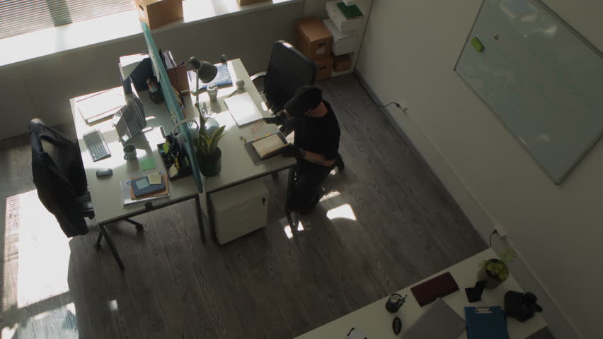 High angle shot of masked man walking into office room searching for confidential corporate data then taking photo of passwords in notebook with mobile phone during industrial espionage