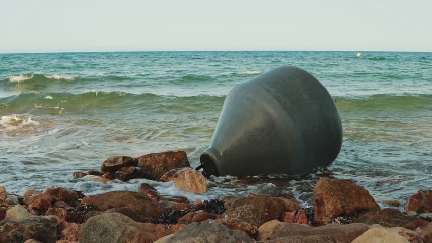 Dark gray buoy gently bobbing against rocky coastline, creating rhythmic movement and serene interaction between maritime navigation element and persistent ocean waves